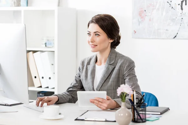 Sonriente atractiva mujer de negocios en traje gris sosteniendo la tableta y utilizando el ordenador en la oficina — Stock Photo