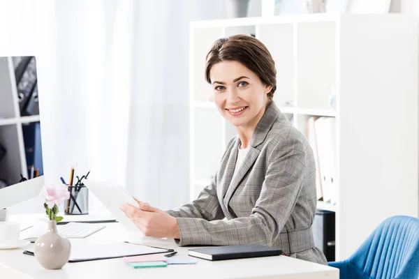 Sonriente atractiva mujer de negocios en traje gris sosteniendo la tableta y mirando a la cámara en la oficina - foto de stock