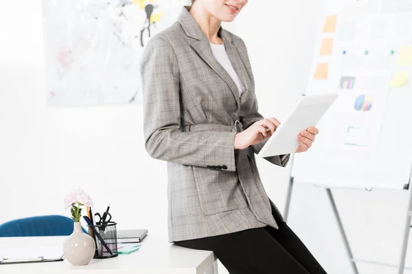 Обрезанный образ деловой женщины в сером костюме, сидящей на столе и использующей планшет в офисе — стоковое фото