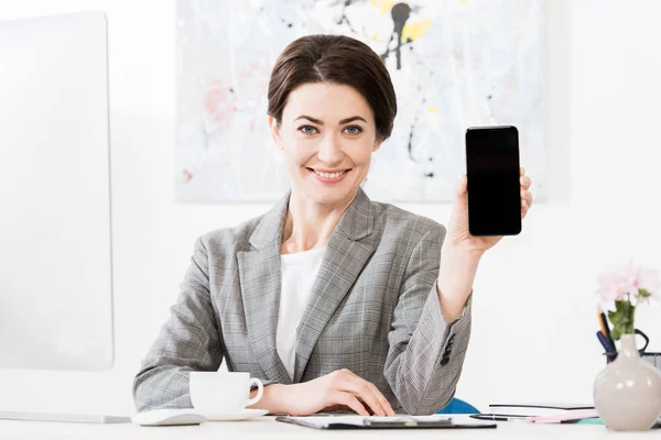 Sonriente atractiva mujer de negocios en traje gris mostrando smartphone con pantalla en blanco en la oficina - foto de stock