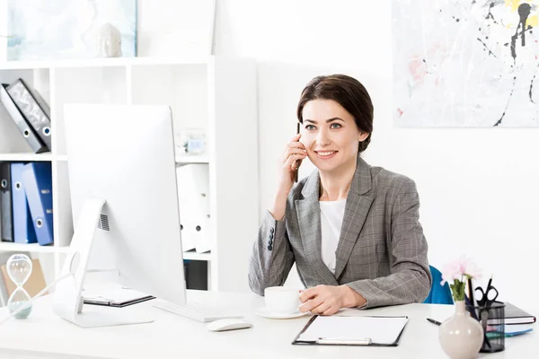 Attractive businesswoman in grey suit talking by smartphone and holding cup of coffee in office — Stock Photo