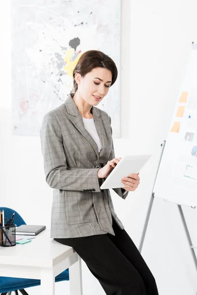 Attractive businesswoman in grey suit sitting on table and using tablet in office — Stock Photo