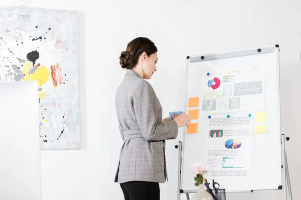 Vue latérale de la femme d'affaires attrayante en costume gris debout près de tableau à feuilles au bureau — Photo de stock