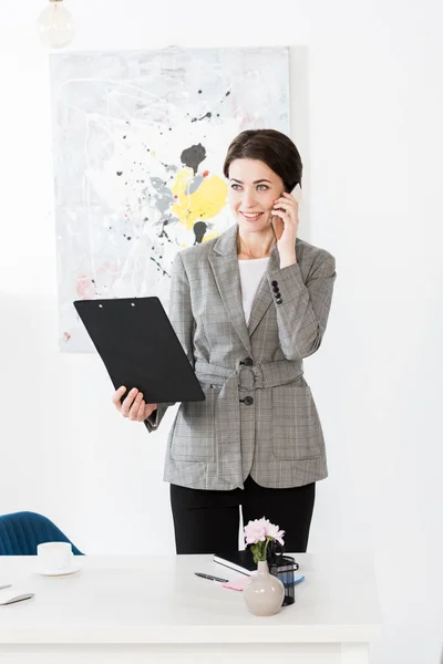Sonriente atractiva mujer de negocios en traje gris hablando por teléfono inteligente y sujetando portapapeles en la oficina - foto de stock