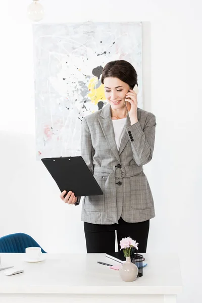 Atractiva mujer de negocios en traje gris hablando por teléfono inteligente y sujetando portapapeles en la oficina - foto de stock