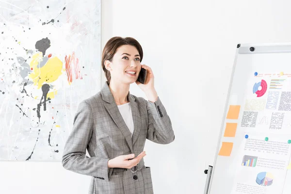 Attractive businesswoman in grey suit standing near flipchart and talking by smartphone in office — Stock Photo