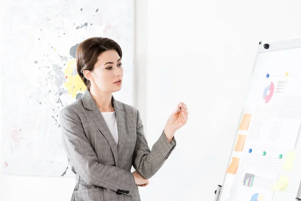 Séduisante femme d'affaires en costume gris regardant le tableau à feuilles mobiles pendant la présentation du projet au bureau — Photo de stock