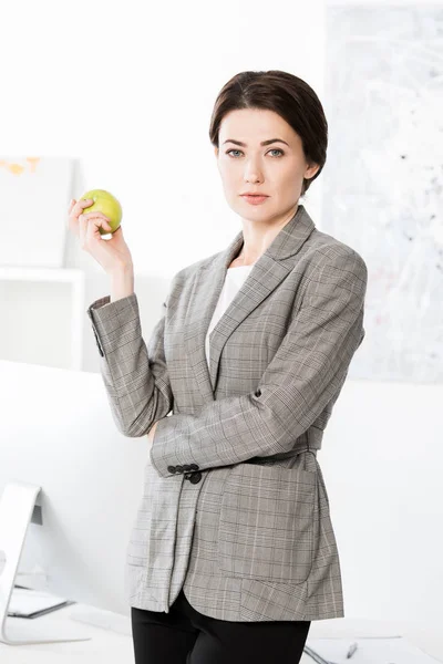Atractiva mujer de negocios en traje gris con manzana y mirando a la cámara en la oficina - foto de stock