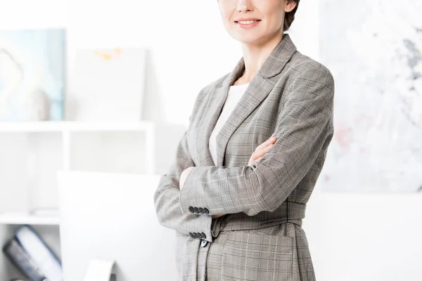 Обрезанный образ деловой женщины в сером костюме, стоящей со скрещенными руками в офисе — стоковое фото
