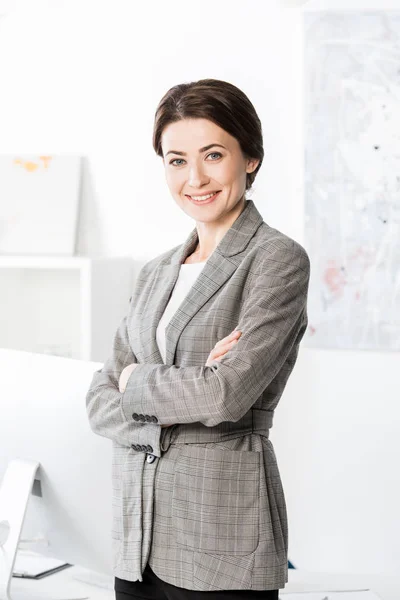 Lächelnde attraktive Geschäftsfrau im grauen Anzug, die mit verschränkten Armen im Büro steht und in die Kamera blickt — Stockfoto