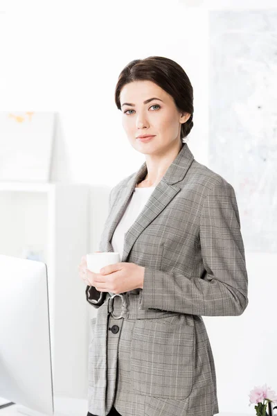 Atractiva mujer de negocios en traje gris sosteniendo taza de café y mirando a la cámara en la oficina - foto de stock