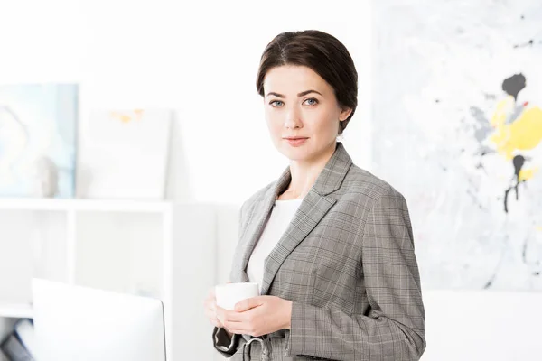 Atractiva mujer de negocios en traje gris sosteniendo taza de té y mirando a la cámara en la oficina - foto de stock