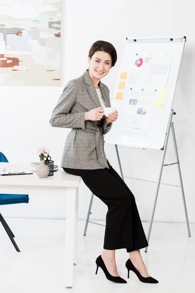 Lächelnde attraktive Geschäftsfrau im grauen Anzug sitzt auf einem Tisch und hält eine Tasse Kaffee im Büro, blickt in die Kamera — Stockfoto