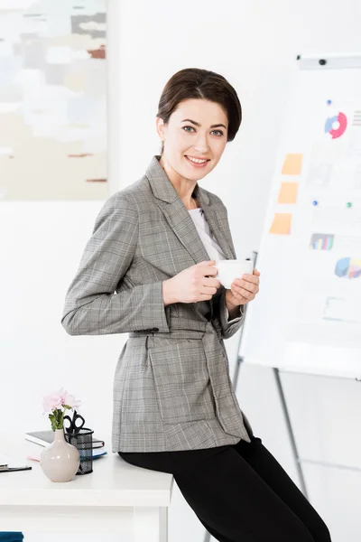 Attraente donna d'affari in abito grigio in possesso di tazza di caffè ans seduto sul tavolo in ufficio — Foto stock