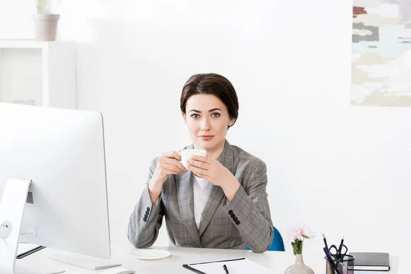 Приваблива бізнес-леді тримає чашку кави за столом і дивиться на камеру в офісі — стокове фото