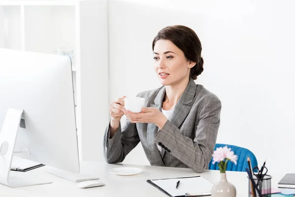 Красивая деловая женщина в сером костюме сидит за столом с компьютером и держит чашку кофе в офисе — стоковое фото
