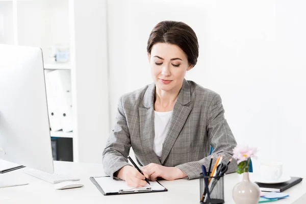 Attractive businesswoman in grey suit writing something to clipboard in office — Stock Photo