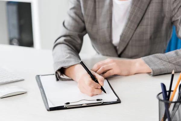 Imagen recortada de la mujer de negocios en traje gris escribiendo algo al portapapeles en la oficina - foto de stock