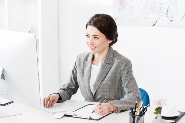 Sonriente atractiva mujer de negocios en traje gris escribiendo algo al portapapeles en la oficina - foto de stock