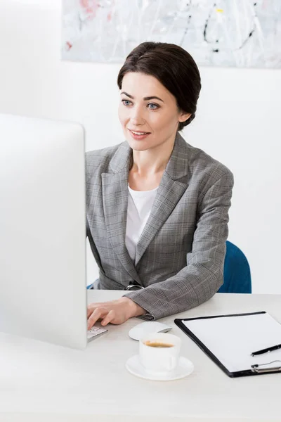 Alegre atractiva mujer de negocios en traje gris sentado usando la computadora en la oficina - foto de stock