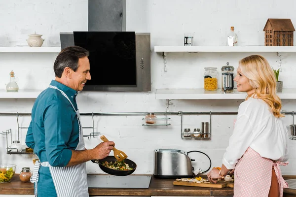 Зрелая жена резки овощей и муж приготовления пищи на сковороде на кухне — стоковое фото