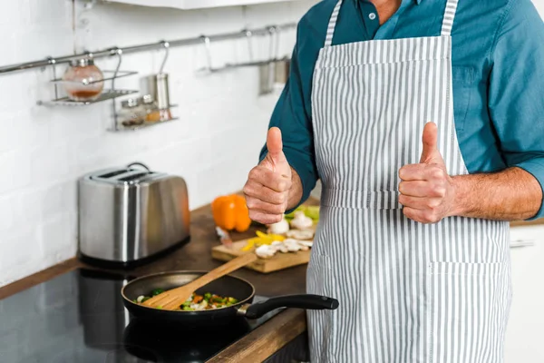 Imagen recortada de hombre maduro cocinar verduras en la sartén en la cocina y mostrando los pulgares hacia arriba - foto de stock