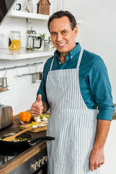 Sonriente guapo maduro hombre cocinar verduras en sartén en cocina y mostrando el pulgar hacia arriba - foto de stock