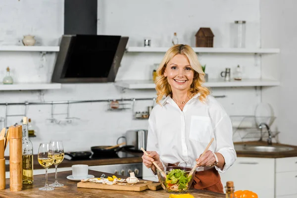 Atractiva mujer madura sonriente mezclando ensalada en la cocina y mirando a la cámara - foto de stock