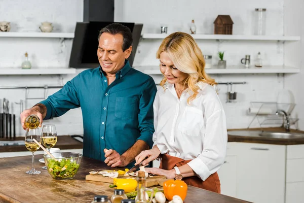 Reife Frau schneidet Gemüse und Ehemann gießt Wein in Gläser in Küche — Stockfoto