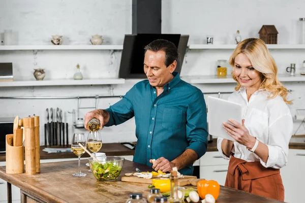 Reife Frau mit Tablette und Mann gießt Wein in Gläser in Küche — Stockfoto