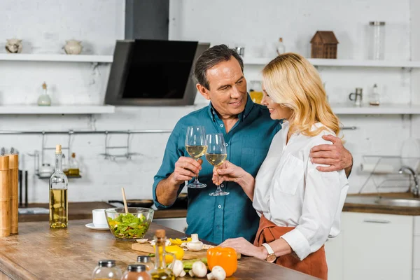 Зрелая жена и муж обнимаются и звенят с бокалами на кухне — стоковое фото