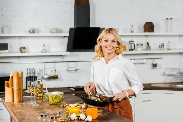 Sonriente atractiva mujer madura sosteniendo sartén con verduras en la cocina y mirando a la cámara - foto de stock