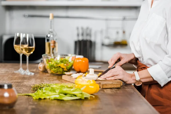 Abgeschnittenes Bild einer reifen Frau, die in der Küche Gemüse auf einem Holzbrett schneidet — Stockfoto