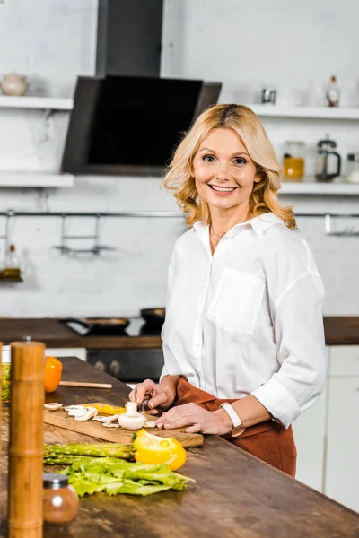 Sonriente atractiva mujer madura cortando verduras en tablero de madera en la cocina y mirando a la cámara - foto de stock