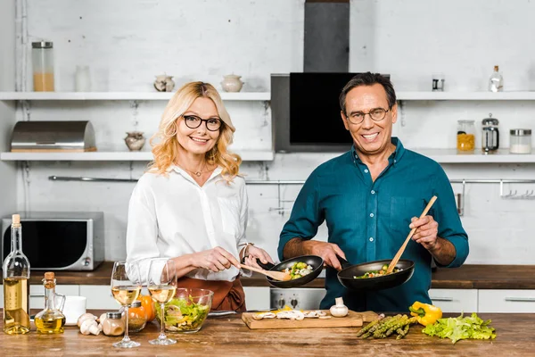 Mature wife and husband cooking and holding frying pans in kitchen — Stock Photo