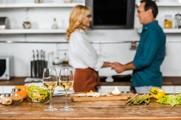 Selektiver Fokus der reifen Frau und des Ehemannes Händchenhalten in Küche, Gemüse und Weingläsern auf der Tischplatte — Stockfoto