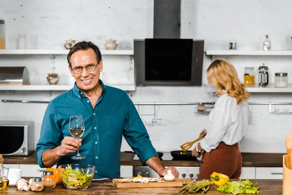 Reife Frau braten Gemüse und Ehemann hält Glas Wein in der Küche — Stockfoto