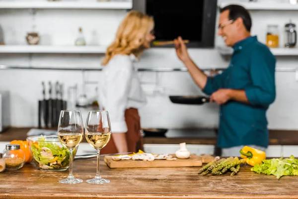 Enfoque selectivo de marido maduro alimentación esposa en cocina, gafas de vino en mesa - foto de stock