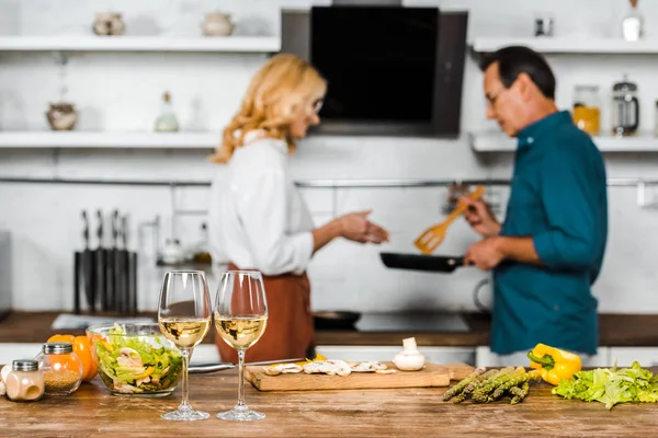 Selective focus of mature husband and wife looking at frying pan in kitchen, wineglasses on tabletop — Stock Photo