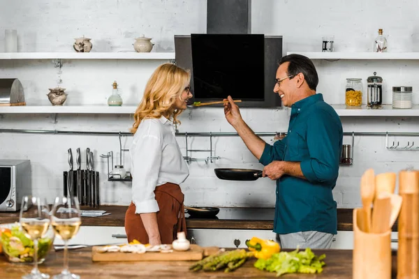 Side view of mature husband feeding wife in kitchen, wineglasses on tabletop — Stock Photo