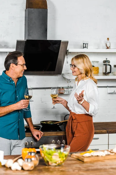 Reife Frau und Mann halten Weingläser in der Hand und schauen einander in der Küche an — Stockfoto