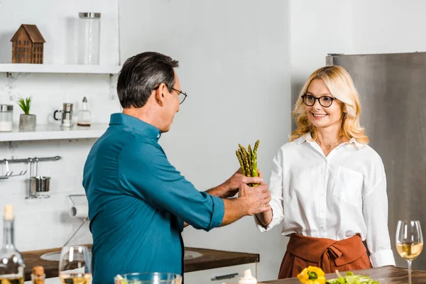 Reife Frau gibt ihrem Mann Spargel zum Kochen in der Küche — Stockfoto