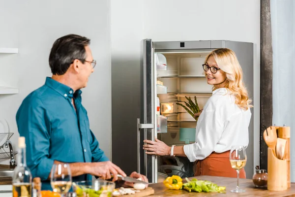 Esposa madura tomando espárragos de la nevera y marido cortando verduras en la cocina - foto de stock