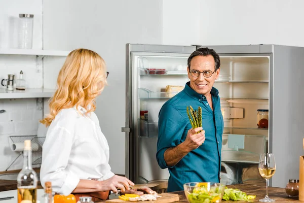 Mature wife cutting vegetables and husband holding asparagus in kitchen — Stock Photo