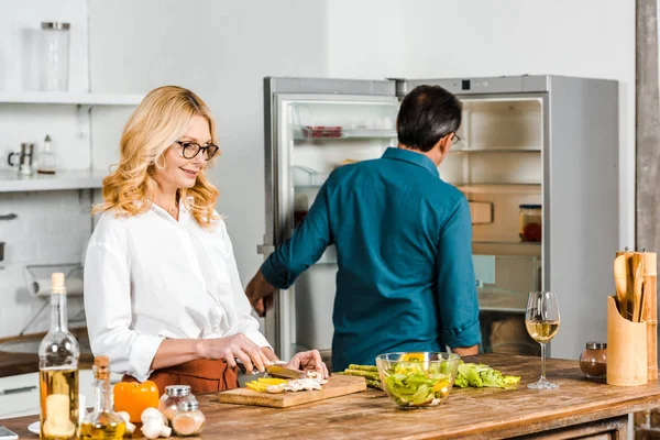 Mature wife cutting vegetables and husband looking at fridge in kitchen — Stock Photo