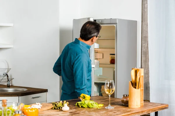 Rückansicht eines reifen Mannes, der in offenen Kühlschrank in der Küche blickt — Stockfoto