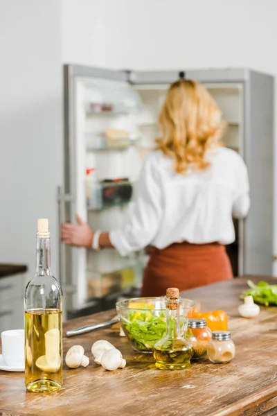Foco seletivo da mulher madura tomando algo da geladeira na cozinha, legumes e vinho no tampo da mesa — Fotografia de Stock