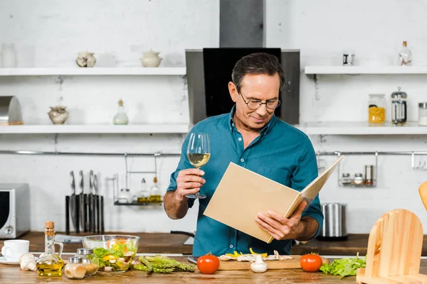 Bel homme mature tenant un verre de vin et livre de cuisine dans la cuisine — Photo de stock