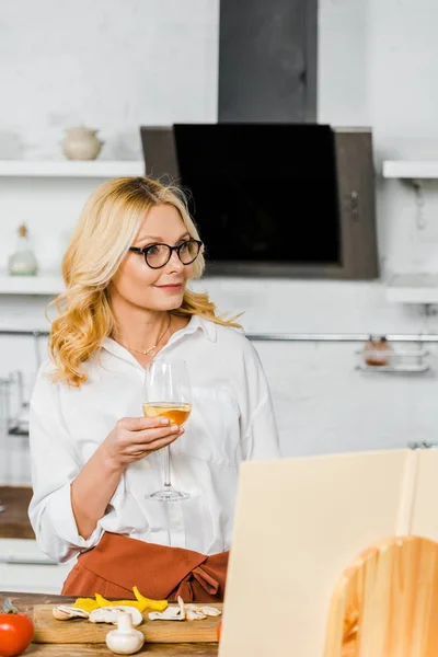Приваблива зріла жінка тримає келих вина і дивиться на кухню — стокове фото