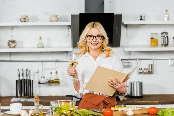 Sorrindo atraente mulher madura segurando copo de vinho e livro de receitas na cozinha — Fotografia de Stock